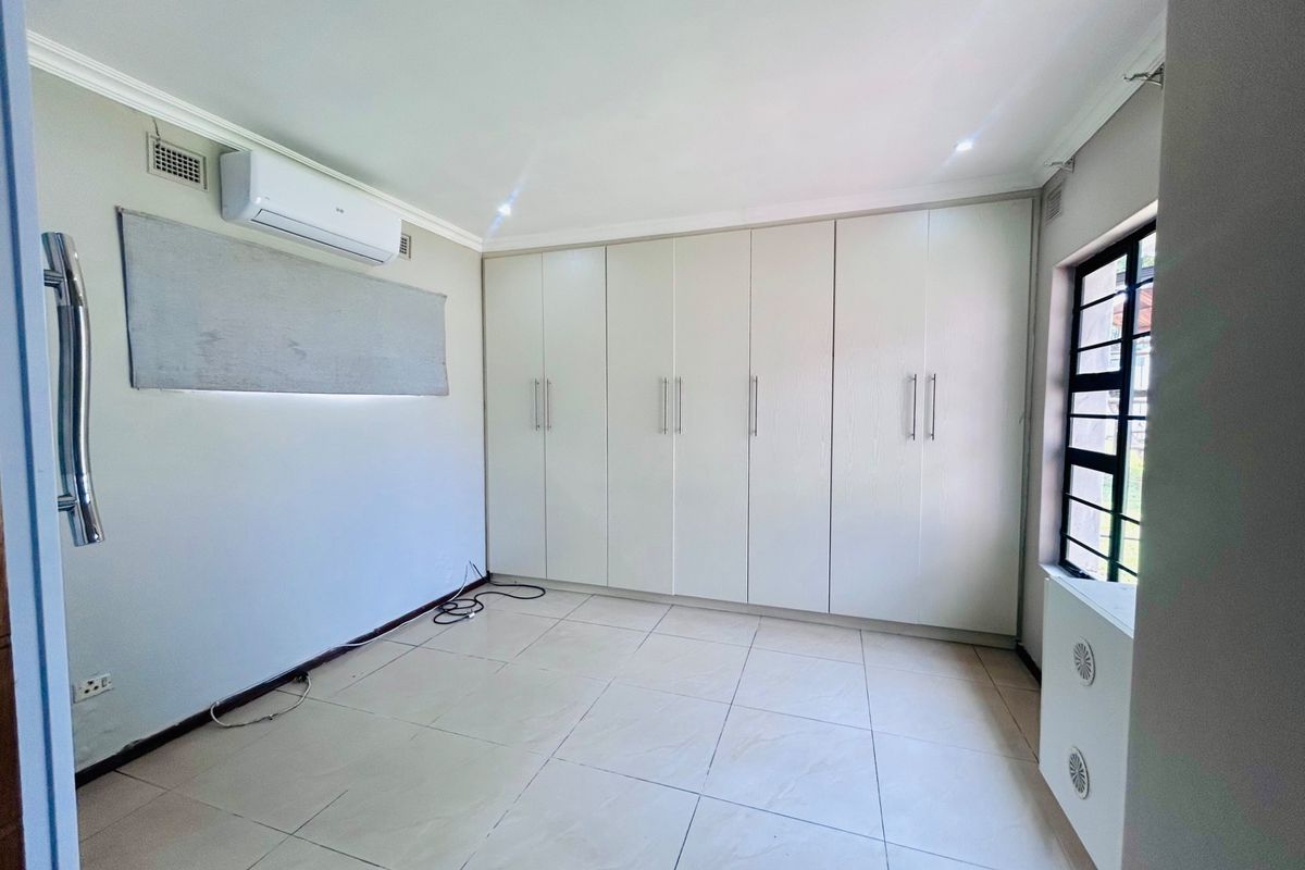A bright, empty bedroom featuring extensive built-in cupboards, tiled flooring, and a wall-mounted air conditioning unit.