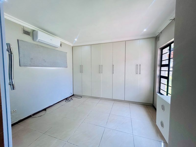 A bright, empty bedroom featuring extensive built-in cupboards, tiled flooring, and a wall-mounted air conditioning unit.