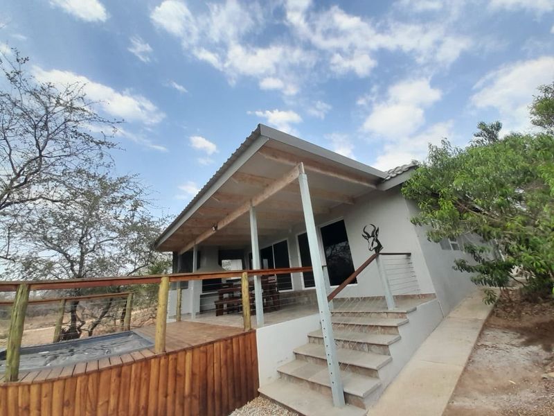 A covered outdoor patio featuring timber decking, a built-in braai, and concrete stairs leading to the entrance of the holiday accommodation.