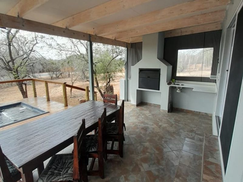 Covered outdoor patio featuring a built-in braai, tiled flooring, exposed timber beams, and an integrated sink, overlooking a natural landscape.