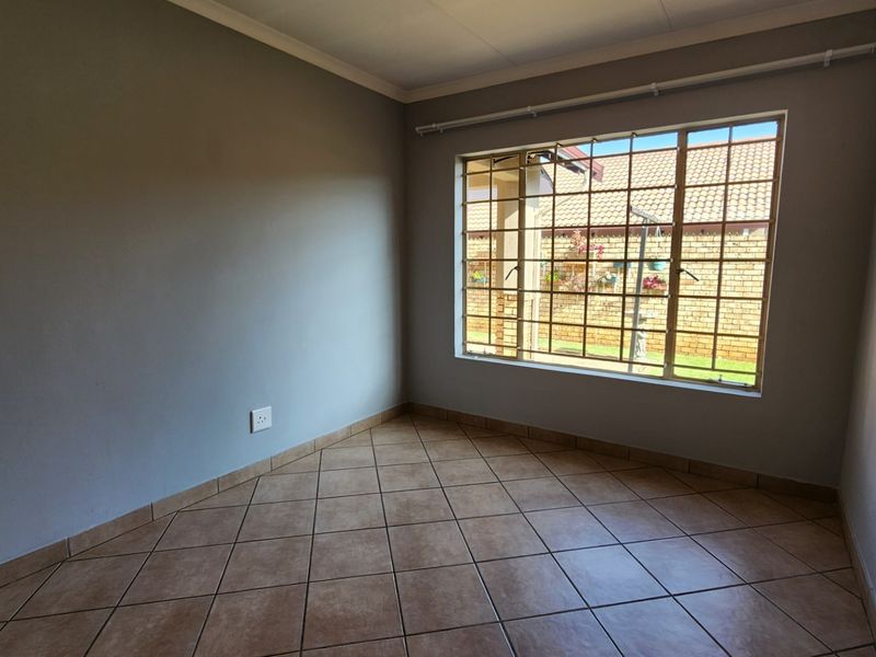 A clean, empty bedroom featuring neutral grey walls, tiled flooring, and a large window fitted with security bars and a curtain rail.