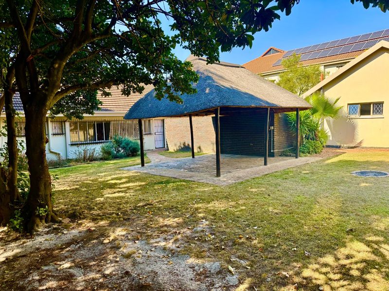 A spacious backyard featuring a traditional thatched roof lapa, paved patio area, and established garden surroundings.