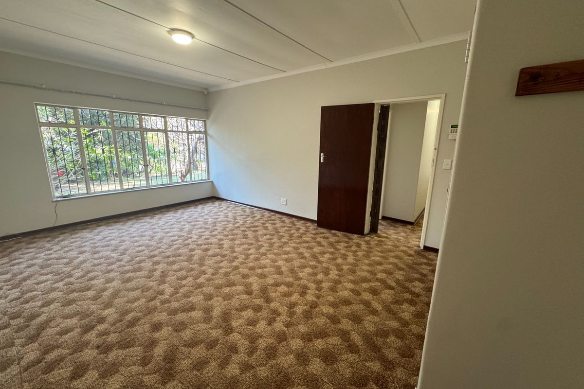 A spacious, empty living room featuring large windows with security bars and dated carpet flooring, offering a blank canvas for renovation.