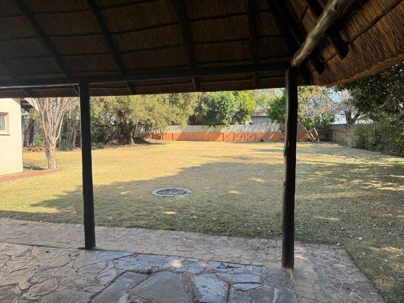 A covered outdoor patio area featuring a traditional thatch roof, timber support posts, and stone paving, overlooking a spacious backyard with a fire pit.