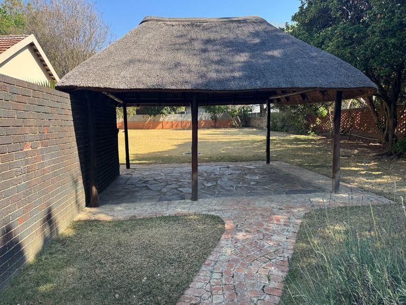An outdoor thatched lapa area featuring stone paving and a brick pathway, set within a spacious garden with boundary walls.