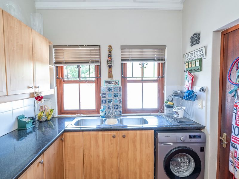 A functional scullery area featuring laminate cabinetry, granite countertops, a double stainless steel sink, and tiled splashback with timber-framed windows.