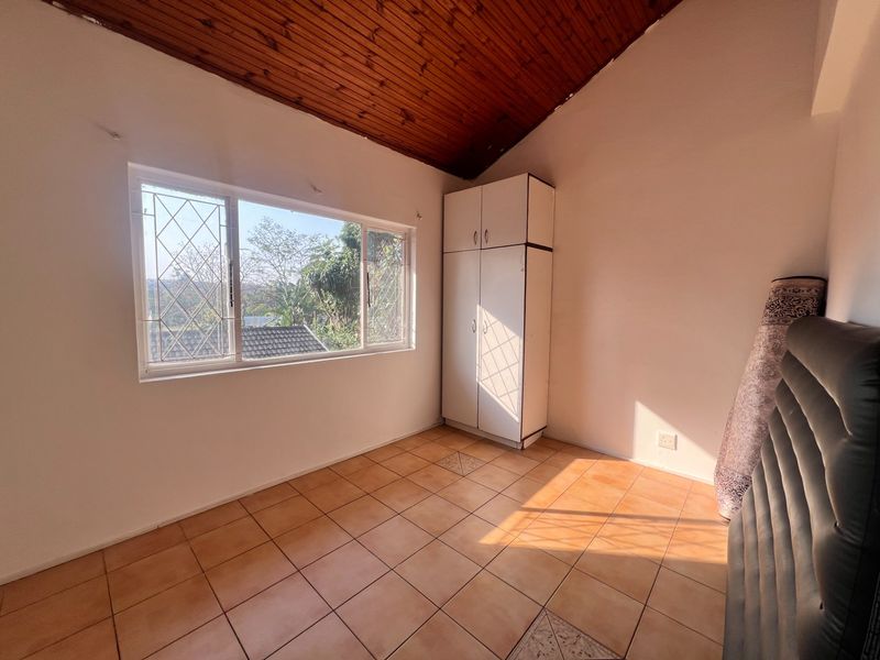 A bright, empty bedroom featuring tiled floors, a timber-clad vaulted ceiling, and a built-in wardrobe with security bars on the window.