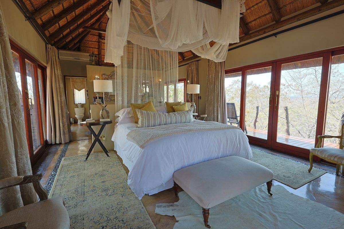 Spacious bedroom featuring a high thatched ceiling with exposed rafters, timber-framed sliding doors opening to a deck, and an integrated en-suite bathroom.
