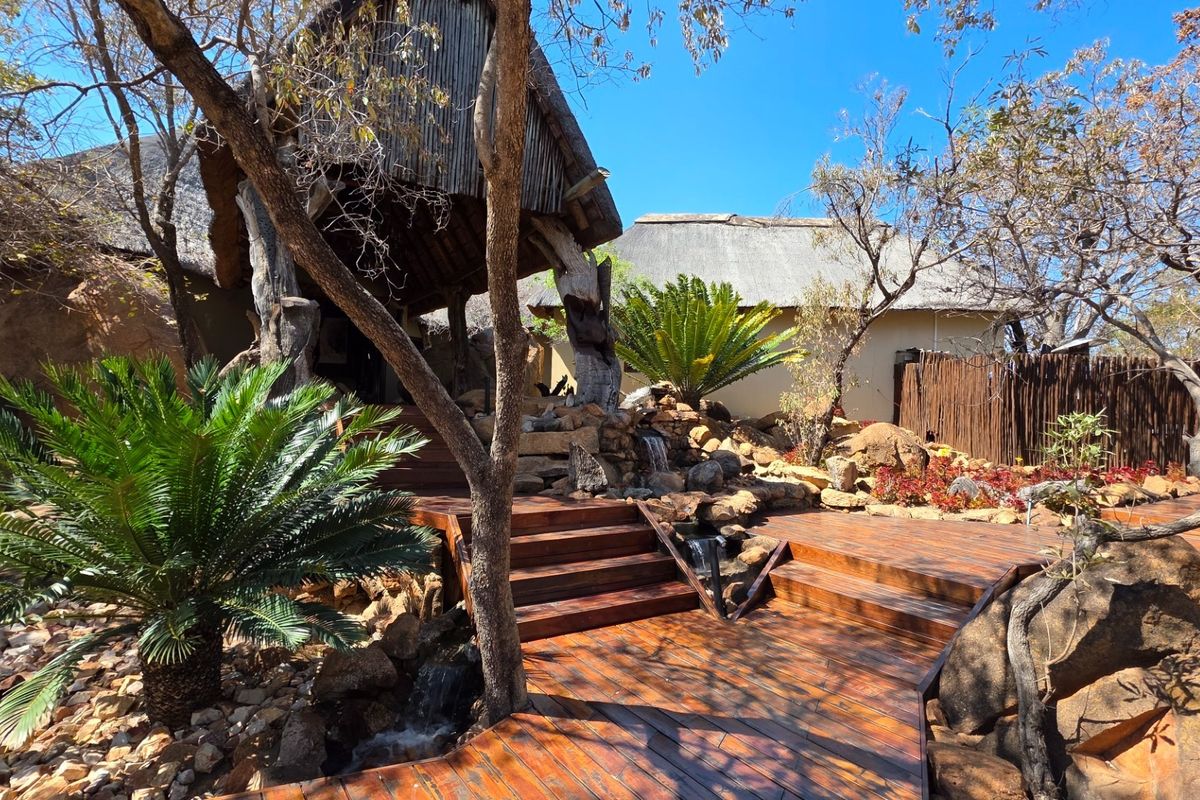 A beautifully landscaped rear garden area featuring extensive timber decking, a natural rockery with a built-in water feature, and traditional thatched roof structures.