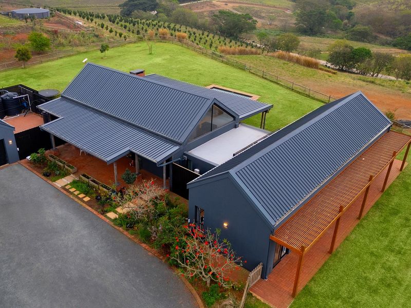Aerial view of a modern farmhouse-style property featuring dark metal roofing, timber pergolas, paved patio areas, and expansive surrounding lawns.