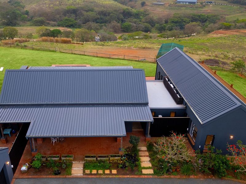 Aerial view of a modern house with a dark metal roof, featuring a spacious covered patio, brick-paved walkways, and a well-maintained garden.