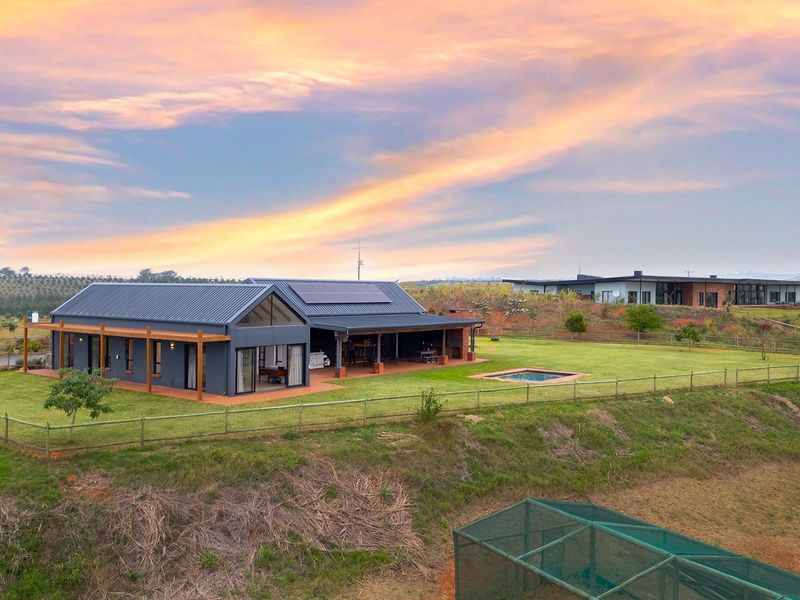 Aerial view of a modern farmhouse with a metal roof, solar panels, covered patio, timber decking, and a swimming pool set in a rural landscape.