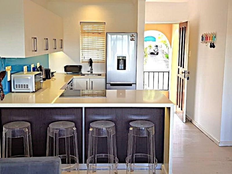 Modern kitchen featuring sleek white cabinetry, stone countertops, a breakfast bar, and a glass splashback, with direct access to an outdoor balcony area.