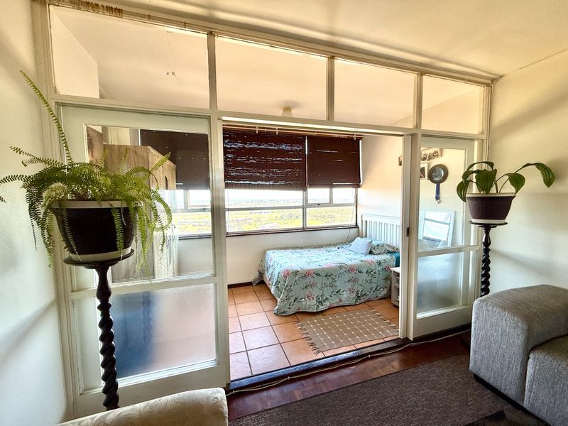 A bedroom featuring terracotta floor tiles and a large window, separated from the living area by a glass partition wall and sliding doors.