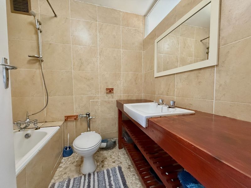 A functional bathroom featuring a bathtub with shower mixer, a toilet, and a wooden vanity with a basin, finished with neutral wall tiles.
