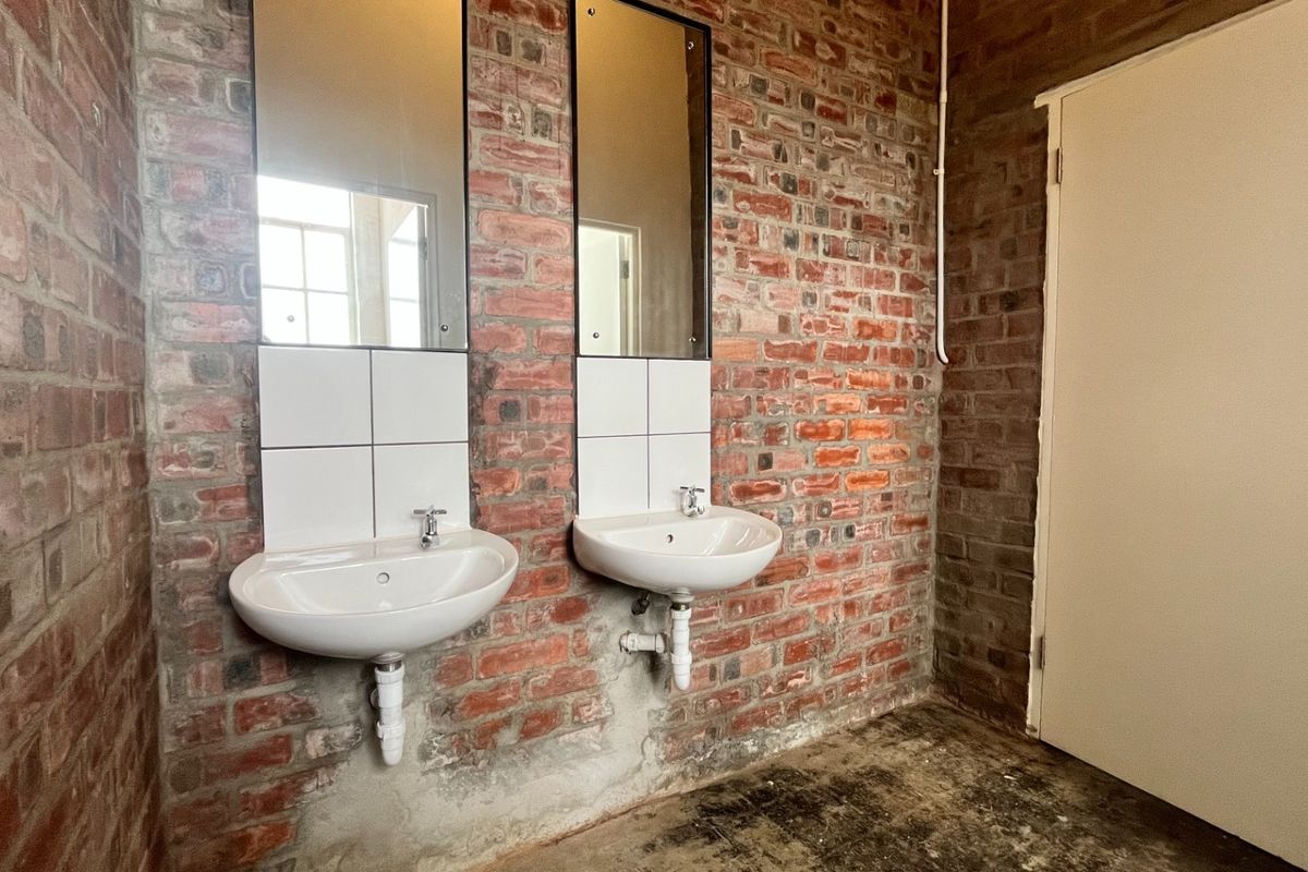 Industrial-style bathroom featuring exposed brick walls, dual wall-mounted basins with chrome fixtures, and simple mirrors, offering a raw, functional aesthetic.