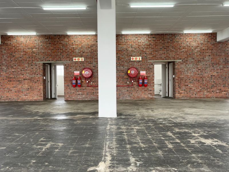 Spacious open-plan industrial unit featuring exposed brick walls, concrete flooring, integrated fire safety equipment, and large steel-framed windows providing ample natural light.