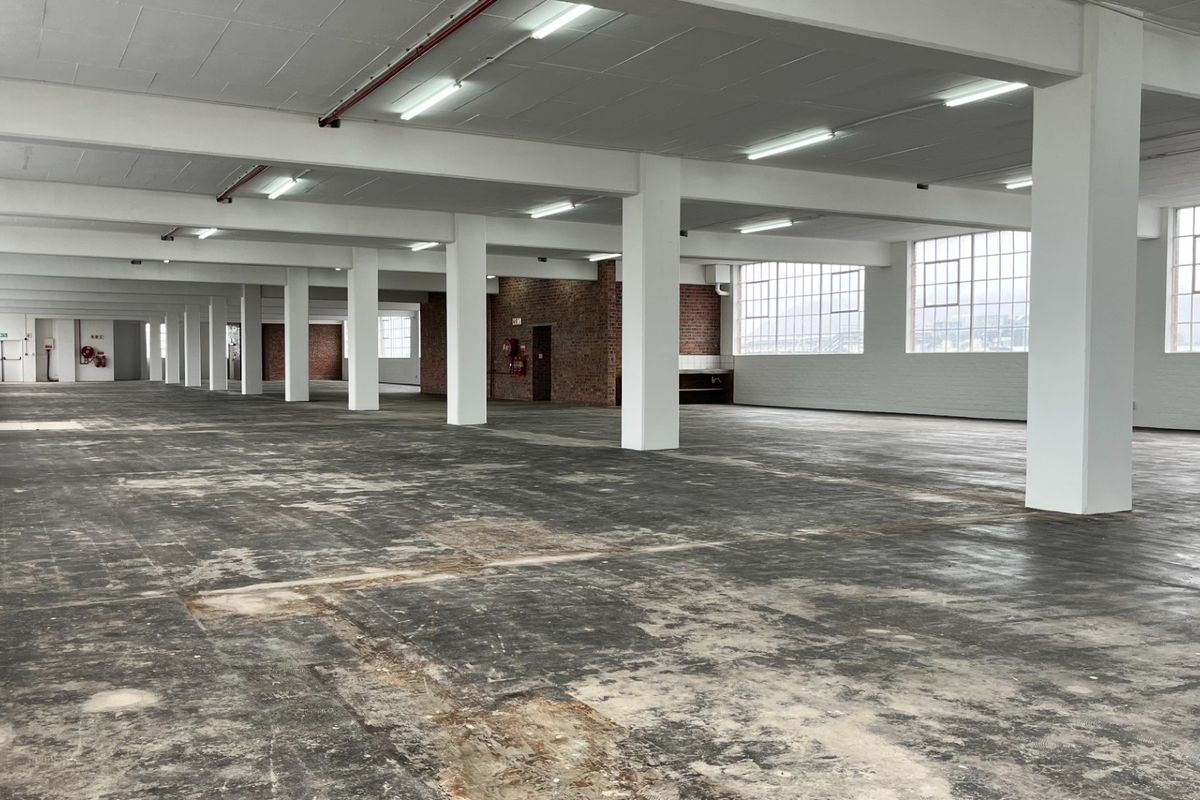 Expansive open-plan industrial warehouse space featuring high ceilings, structural columns, exposed brick accents, and large steel-framed windows providing ample natural light.