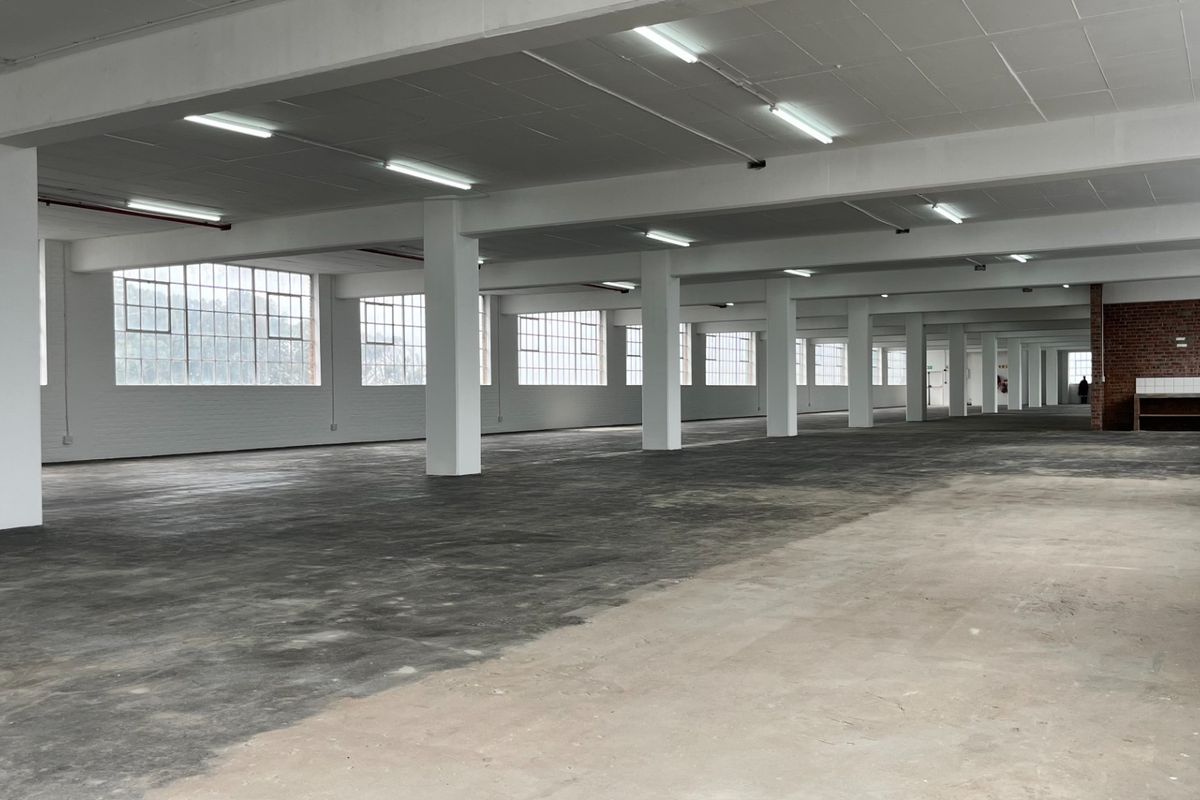 Expansive open-plan industrial warehouse space featuring high ceilings, abundant natural light from steel-framed windows, concrete flooring, and a dedicated sink area.