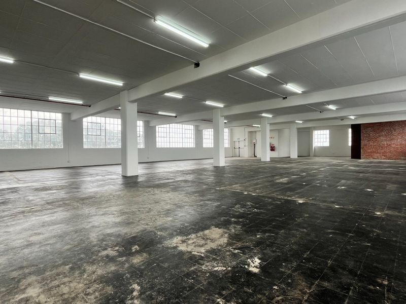 Expansive open-plan industrial warehouse space featuring large steel-framed windows, exposed structural columns, and a distinctive brick feature wall.