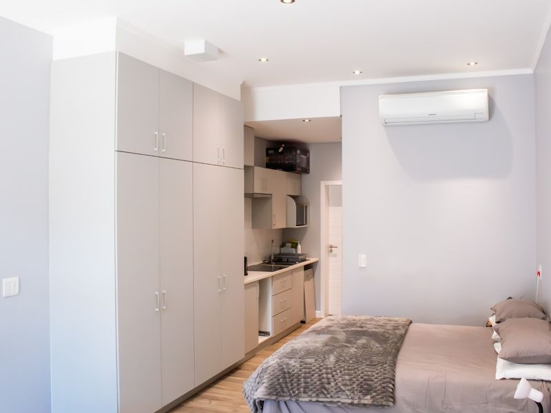 A modern bedroom featuring built-in cupboards, a compact kitchenette with sink and extractor, and a wall-mounted air conditioning unit.