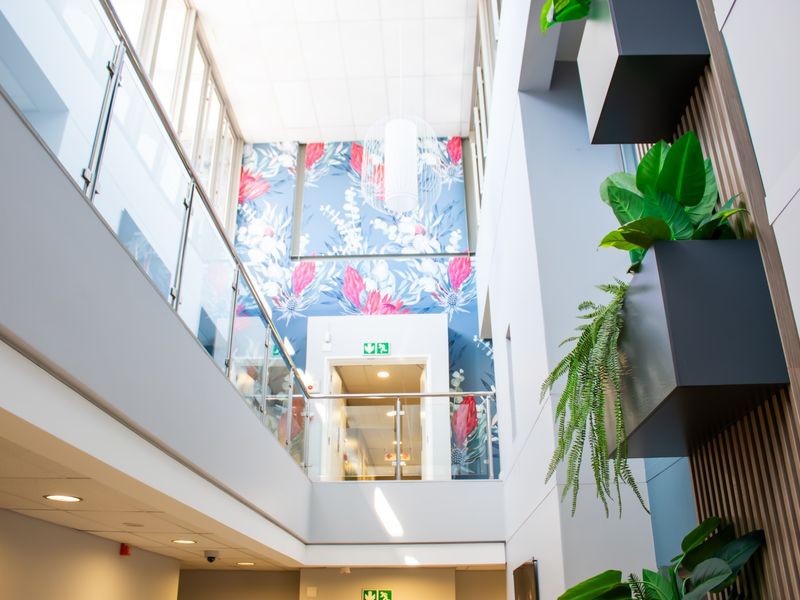 A bright, modern atrium-style hallway featuring a glass balustrade, high ceilings, and decorative floral feature wall in a well-maintained retirement village facility.