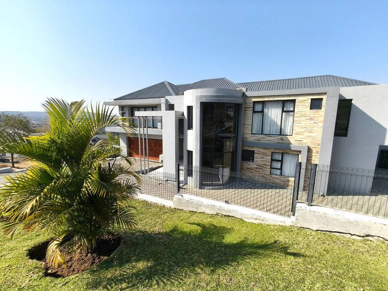 Modern double-storey residence featuring a striking curved glass facade, stone cladding accents, metal roofing, and a secure gated driveway entrance.