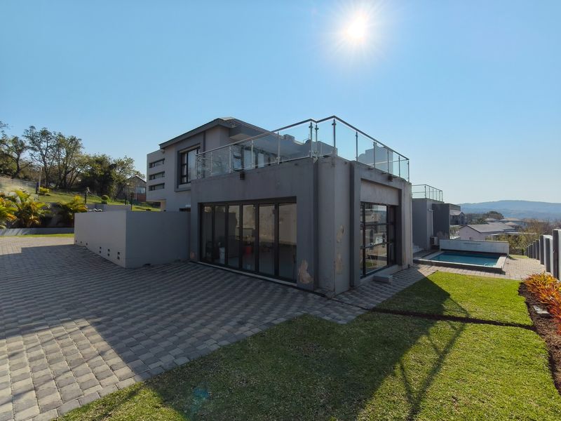 Rear view of a modern multi-level home featuring a glass balustrade balcony, paved patio area, swimming pool, and landscaped garden.