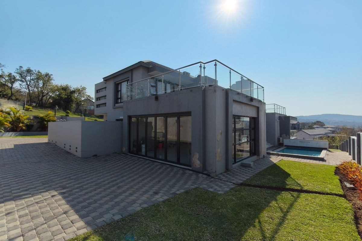 Rear view of a modern multi-level home featuring a glass balustrade balcony, paved patio area, swimming pool, and landscaped garden.