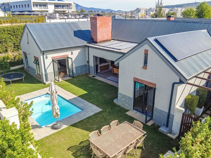Aerial view of a residential property featuring a swimming pool, landscaped garden, solar panels, and a covered patio area with a built-in braai.