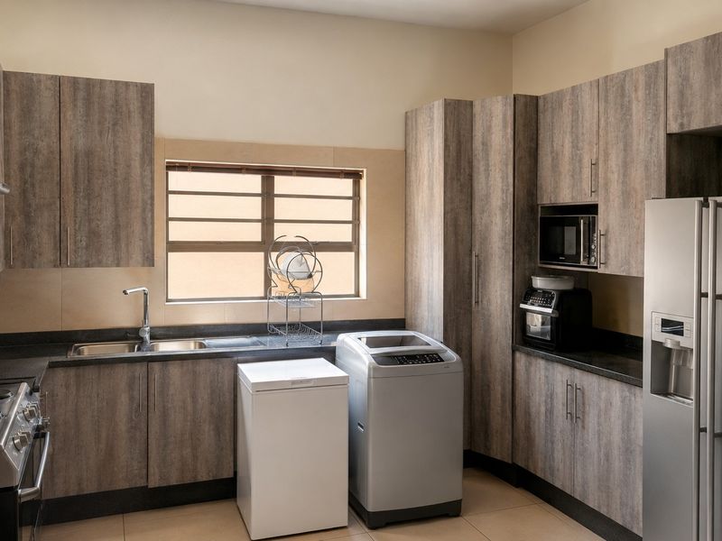 A functional scullery featuring modern wood-look cabinetry, stone countertops, integrated appliances, and a stainless steel sink with a window for natural light.