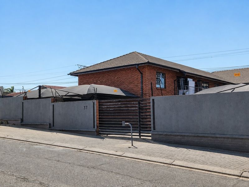 Front exterior view of a brick house featuring a secure concrete boundary wall, electric fencing, and a metal driveway gate.