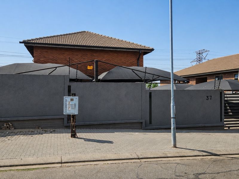 Front exterior view of the property featuring a secure perimeter wall with electric fencing, paved driveway, and covered carport structures.