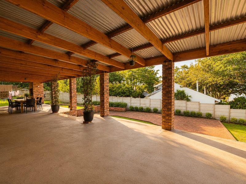 Spacious covered patio featuring exposed timber beams, corrugated metal roofing, and brick columns, overlooking a landscaped garden with paved pathways.