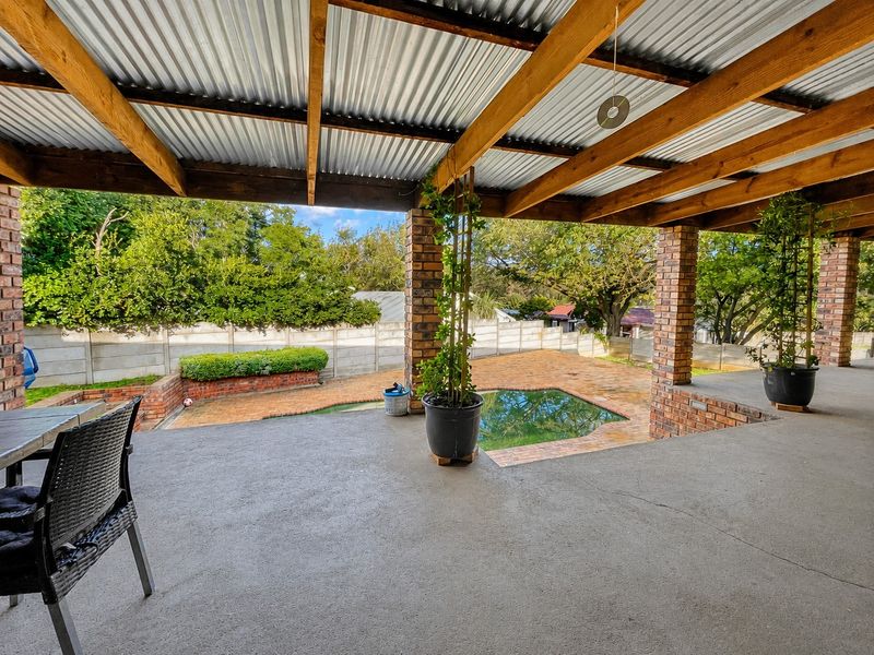 Covered outdoor patio featuring exposed timber beams and brick pillars, overlooking a swimming pool and paved garden area with boundary walling.