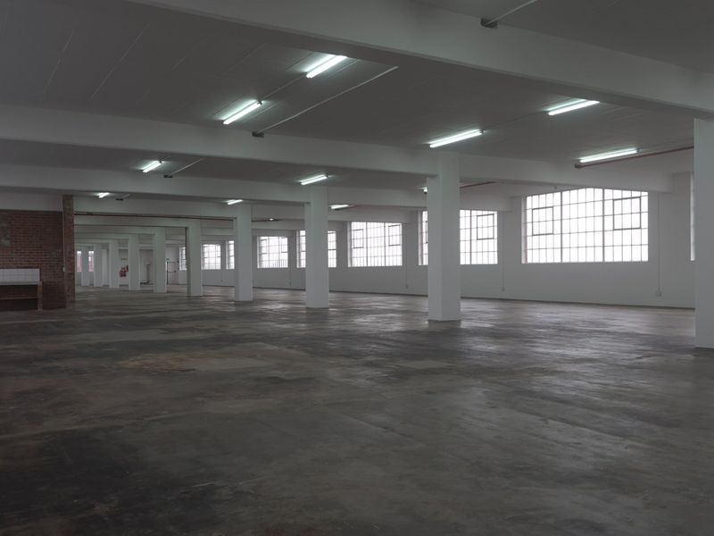 A large, open-plan industrial warehouse space featuring concrete flooring, exposed brick accents, and ample natural light through large steel-framed windows.