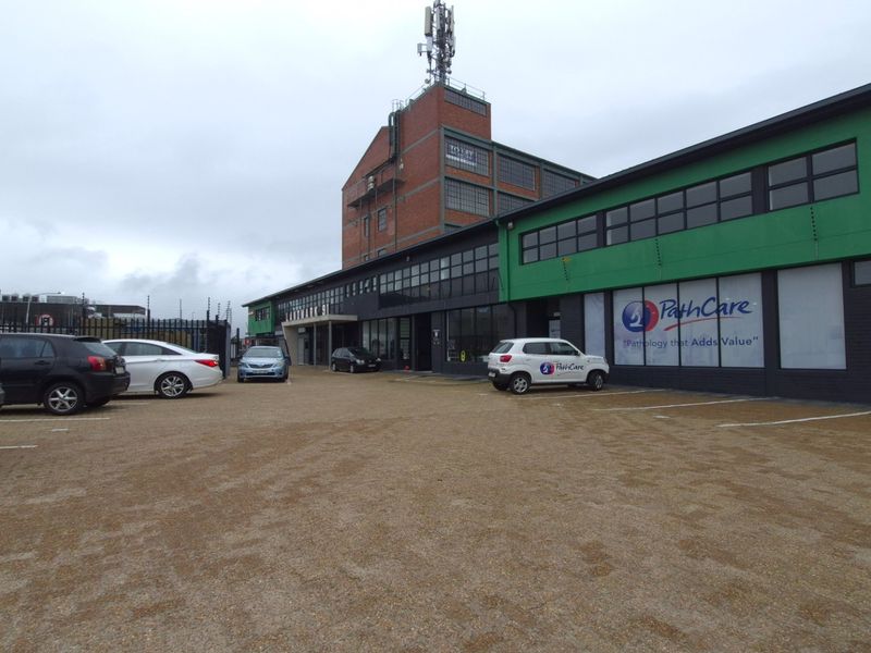 Exterior view of a multi-story industrial property featuring a large paved parking area, modern facade, and secure perimeter fencing.