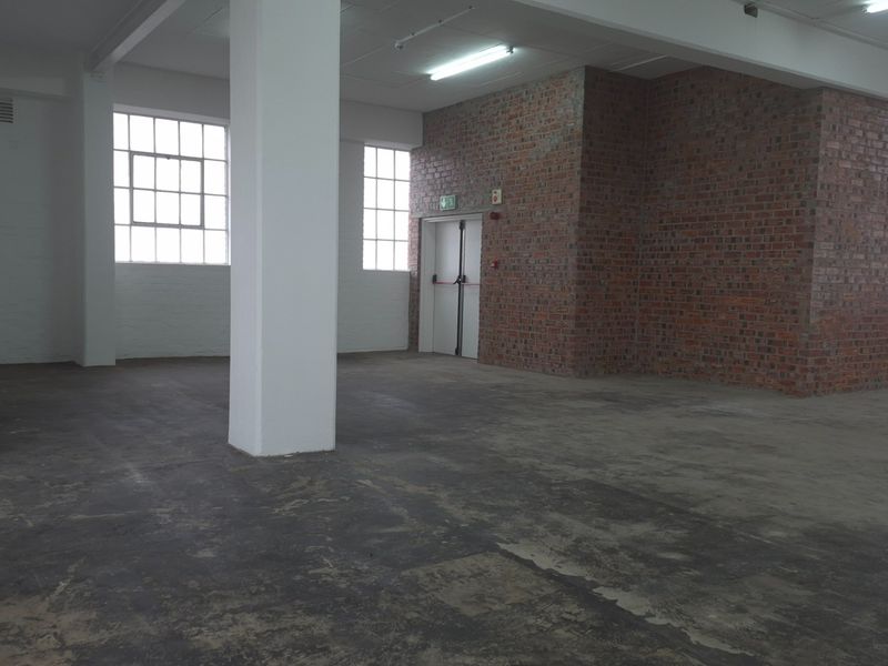 Spacious open-plan industrial unit featuring exposed brickwork, concrete flooring, large steel-framed windows, and functional overhead lighting, suitable for various commercial or creative uses.