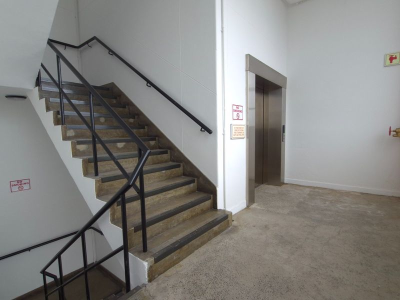 Industrial hallway featuring a concrete staircase with metal handrails, a functional elevator, and exposed fire safety infrastructure.