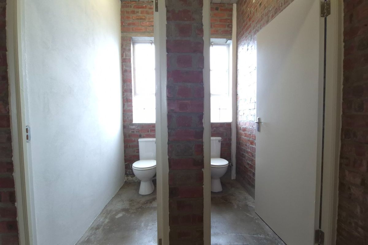 Two separate toilet cubicles featuring exposed brick walls, concrete flooring, and natural light from windows.