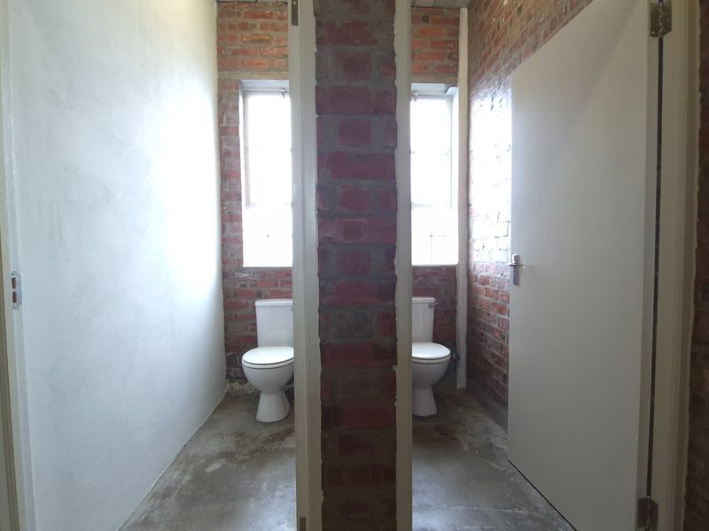 Two separate toilet cubicles featuring exposed brick walls, concrete flooring, and natural light from windows.