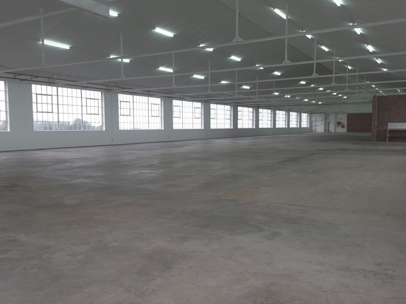 Spacious industrial warehouse area featuring high ceilings, extensive natural light through large steel-framed windows, concrete flooring, and a dedicated utility counter.
