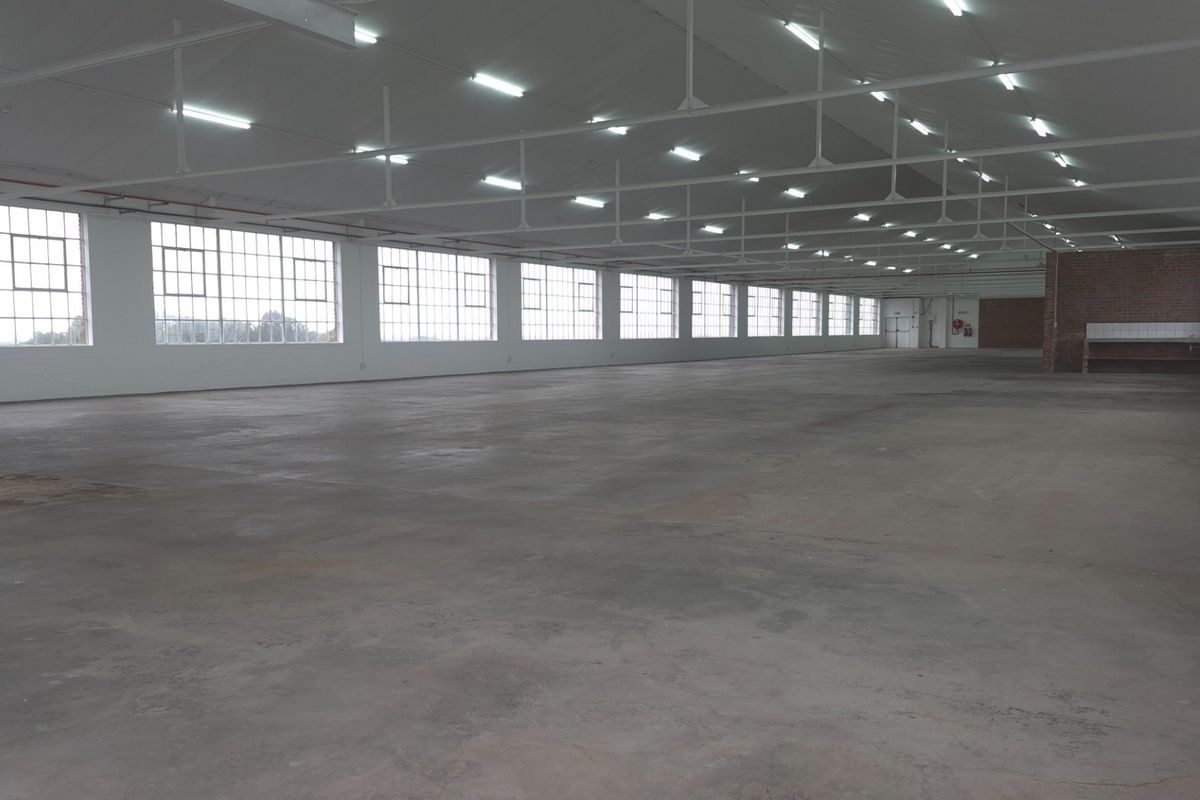 Spacious industrial warehouse area featuring high ceilings, extensive natural light through large steel-framed windows, concrete flooring, and a dedicated utility counter.