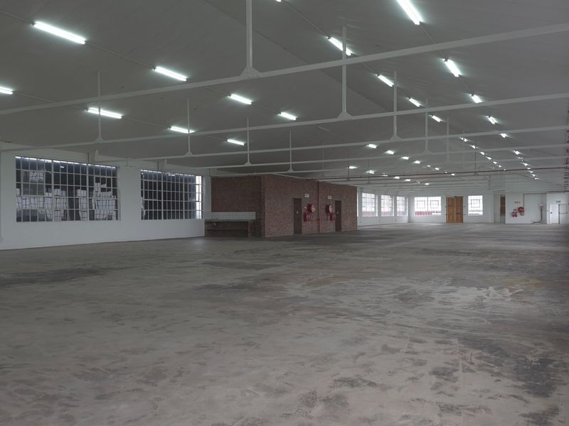 Expansive industrial warehouse space featuring high ceilings with exposed steel trusses, large windows for natural light, and durable concrete flooring.