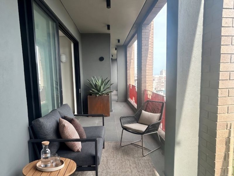 A modern covered balcony featuring tiled flooring, brick structural pillars, and sliding glass door access, offering a clean and well-maintained outdoor living space.