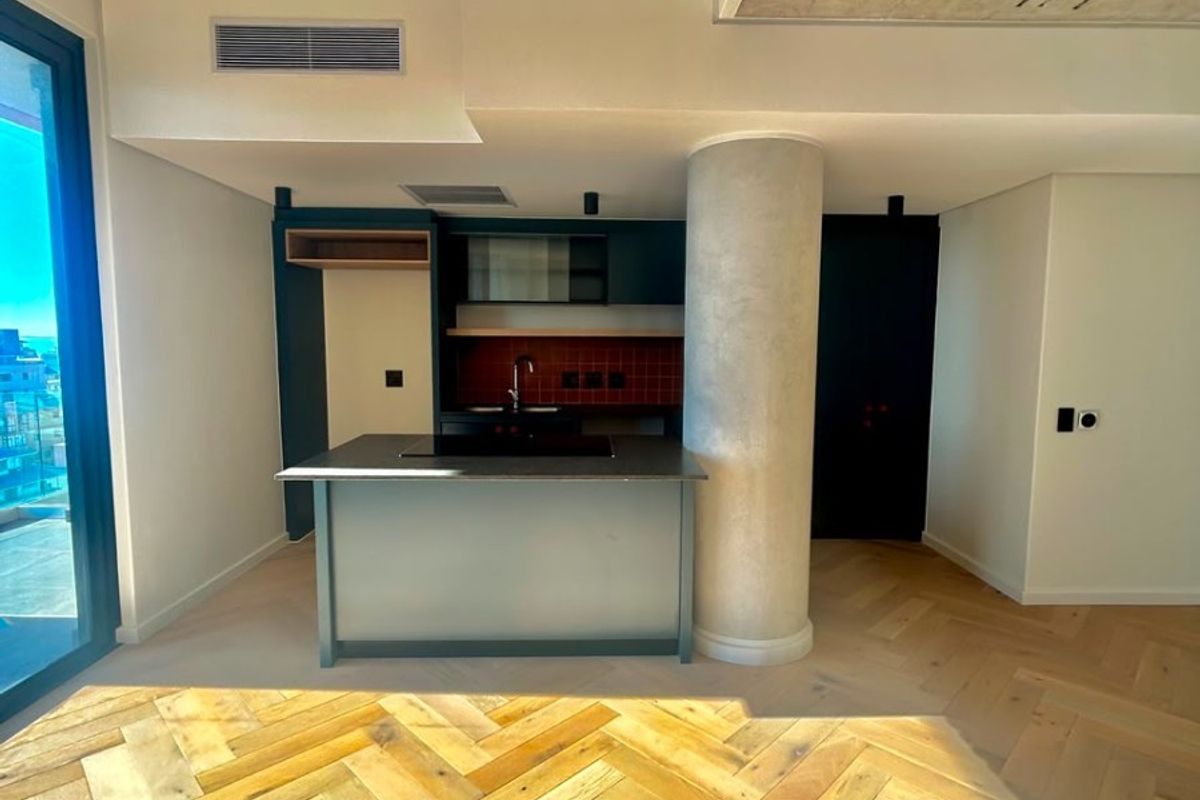 Modern kitchen area featuring a central island, built-in hob, sink, herringbone timber flooring, and an exposed concrete ceiling in a contemporary open-plan space.
