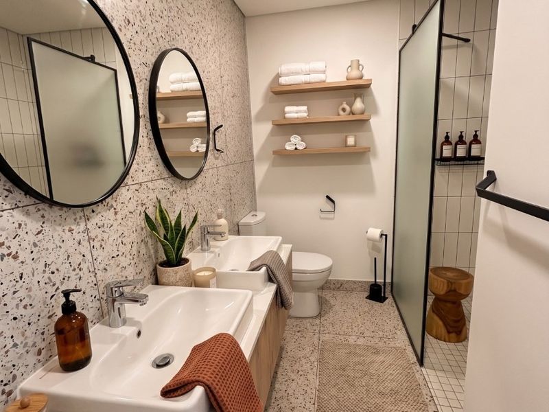 Modern bathroom featuring a double vanity with terrazzo wall tiling, floating timber shelving, and a spacious walk-in shower with a glass screen.