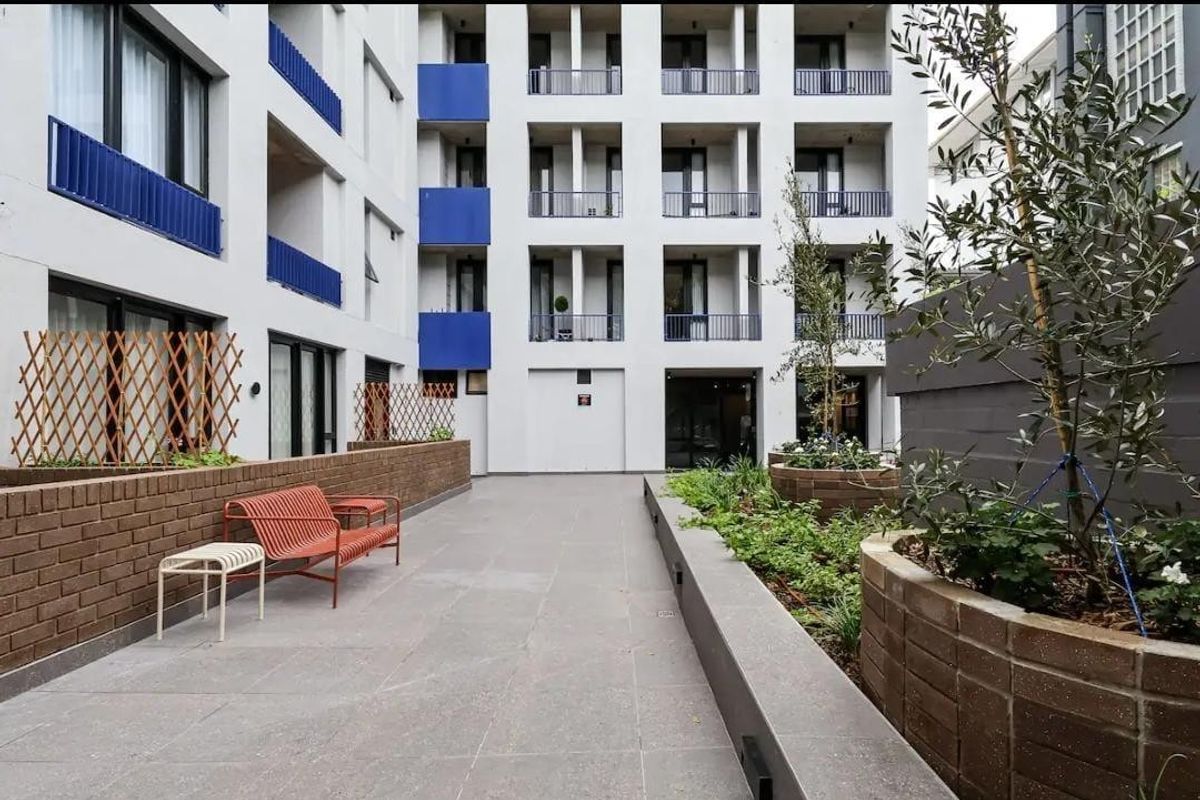 A modern, well-maintained apartment complex courtyard featuring tiled flooring, integrated brick planter boxes, and private balconies with blue metal railings.