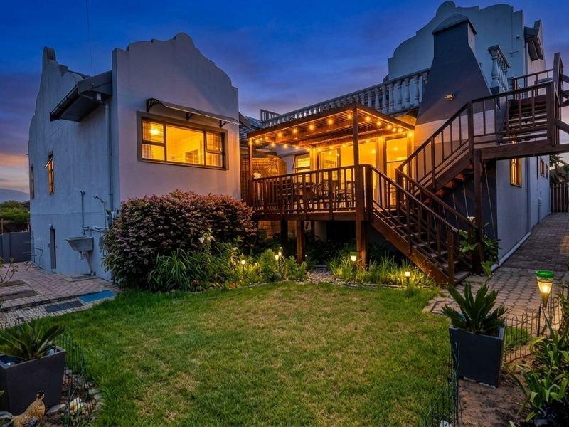 A beautifully illuminated rear exterior featuring a timber deck with a pergola, external staircase, and a well-maintained garden area at twilight.