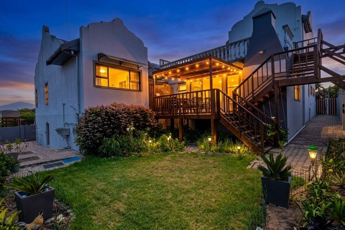 A beautifully illuminated rear exterior featuring a timber deck with a pergola, external staircase, and a well-maintained garden area at twilight.